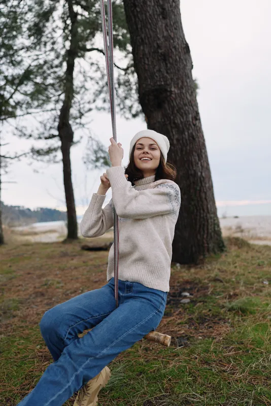 Woman on swing outdoors among trees, joyful casual style, wearing a sweater and beanie, enjoying nature with a relaxed confident smile in daylight and fresh air.
