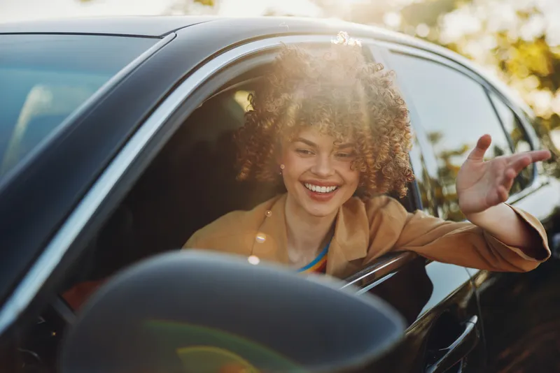 A smiling young woman leans out of a car window during golden hour, retro film color adding warmth and a candid street vibe, suggesting carefree summer travel.