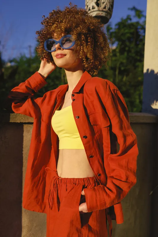 Cheerful woman outdoors in a bright yellow top and red jacket, sunlit portrait, relaxed pose, curly hair, stylish casual lifestyle, warm colors, natural backdrop