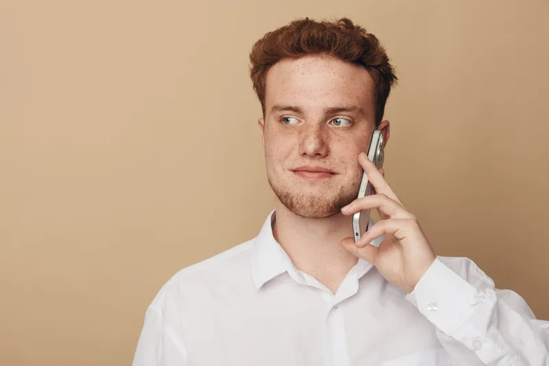 Young man with red hair and beard wearing white shirt talks on smartphone looking away on beige background. Concept of communication, technology, and casual lifestyle.