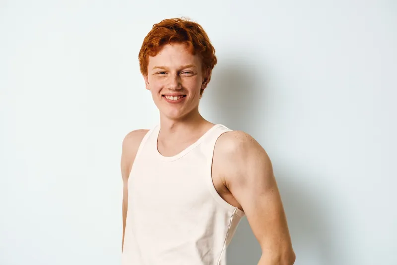 Young man with red hair wearing white tank top smiling and standing confidently against plain white background. Casual portrait with natural expression and relaxed posture.