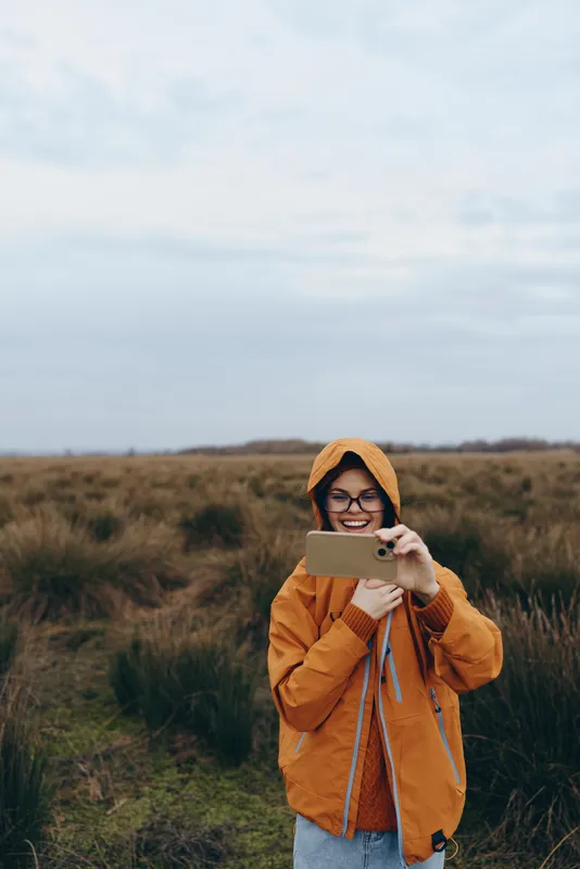 Woman lifestyle outdoors, with a smile taking a photo with a smartphone, wearing orange jacket, knit hat, glasses, standing in a grassy field, soft daylight, travel mood, autumn nature