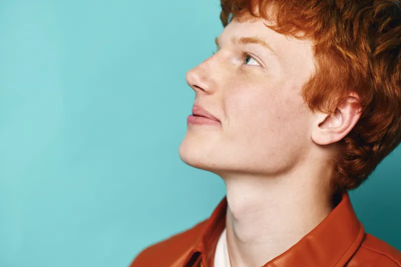 Portrait of young redhead man looking up with thoughtful expression on blue background. Casual clothing and soft lighting highlight skin texture and hair details in closeup profile view.