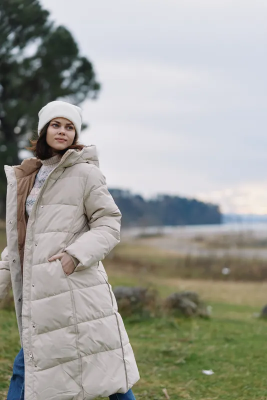 Winter coat worn by a woman in an outdoor landscape, beige puffer jacket, beanie, calm shoreline in the background, casual stylish cold weather look