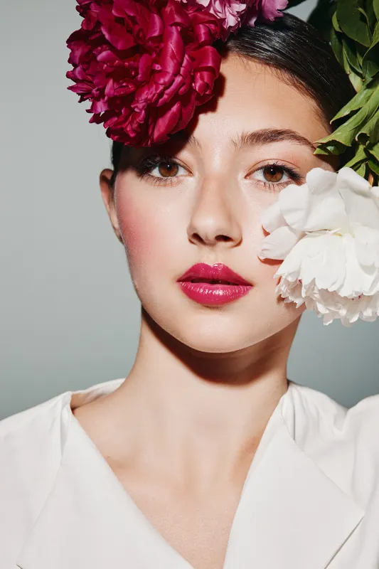 Young woman with fresh natural makeup poses with vibrant red and white flowers by face. Clean background highlights bracelets of creative beauty and fashion concept.