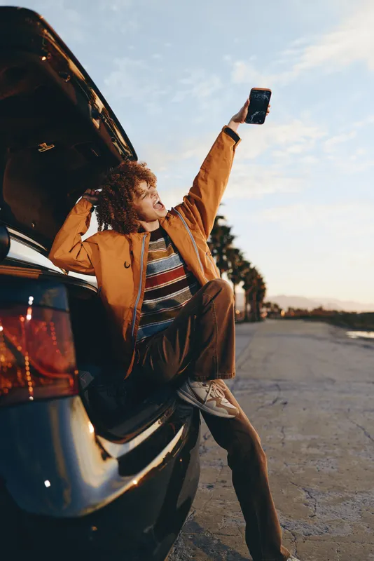 Woman lifestyle outdoors, standing by car on roadside, raising bottle toward sky at sunset, curly hair, casual jacket, jeans, relaxed smile, warm light, summer travel mood, golden glow