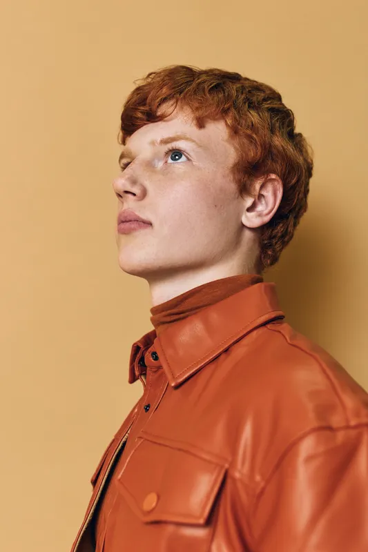 Young man with red hair wearing brown leather jacket looking sideways on beige background. Portrait of confident male model with clear skin and sharp facial features in studio.