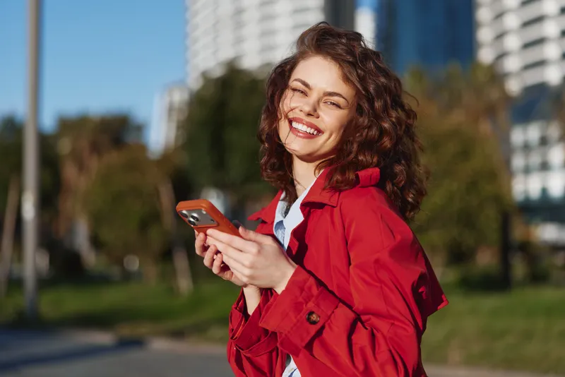 Smiling young woman with curly brown hair wearing a red jacket holds a smartphone in an outdoor park with modern buildings in the background on a sunny day.