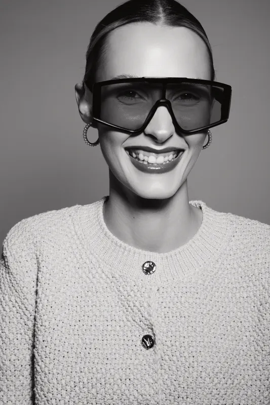 Smiling Caucasian woman with pale skin wearing large sunglasses and textured sweater in studio against gray background.