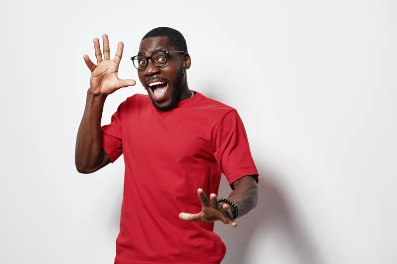 African American man with dark skin wearing glasses and red t shirt showing five fingers with excited expression on white background. Casual style and positive gesture for counting or greeting.