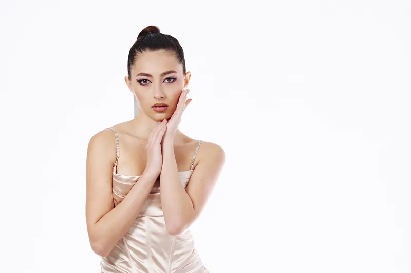 Elegant Asian woman in satin dress delicately touches her face. Soft makeup and sleek bun for beauty, fashion and creative portrait on white background.