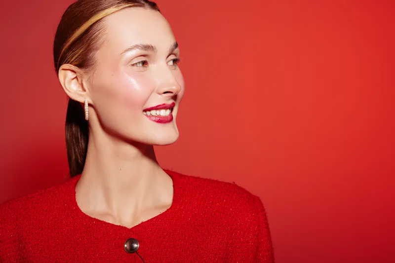 Smiling woman with glossy skin and red lipstick looks sideways on red background. She wears red clothing and gold earrings. Concept of beauty, fashion, makeup, and elegance in studio.
