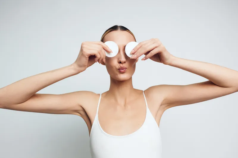 A young woman in a tank top performs a home facial, pressing cotton pads over her eyes in a bright studio to convey self care, relaxation, skincare, and a calm moment.