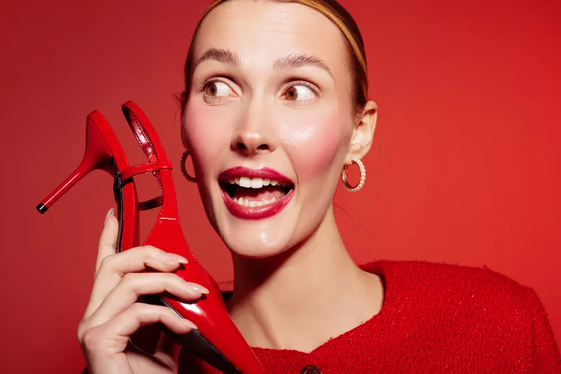 Young woman with red lipstick and hoop earrings holds a red high heel shoe near her face, smiling and looking sideways on a red background. Concept of fashion, beauty, and style.
