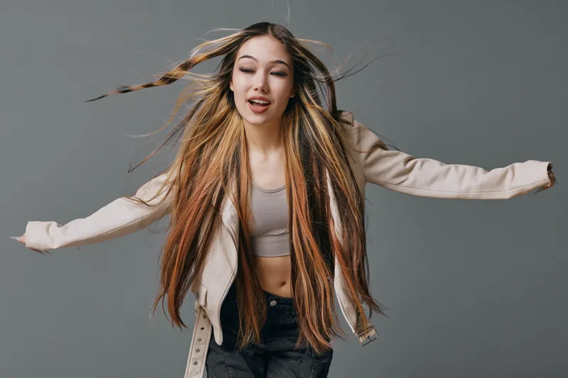Young woman with long braided hair and casual clothes dancing and joyfully moving with arms stretched out over gray background in studio. Happy girl with motion in hair.