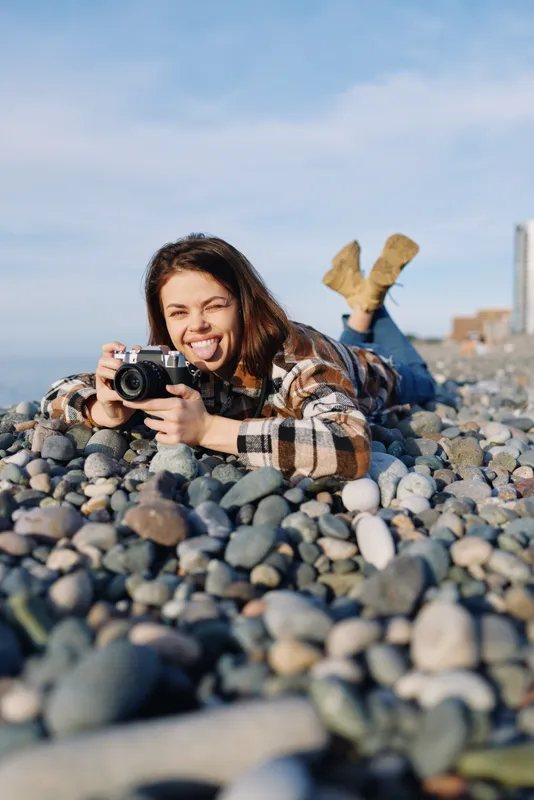 Woman photographer lying on a rocky beach, aiming a camera with focus and joy. Outdoor scene captures candid creativity, natural light, and relaxed mood in a coastal setting.
