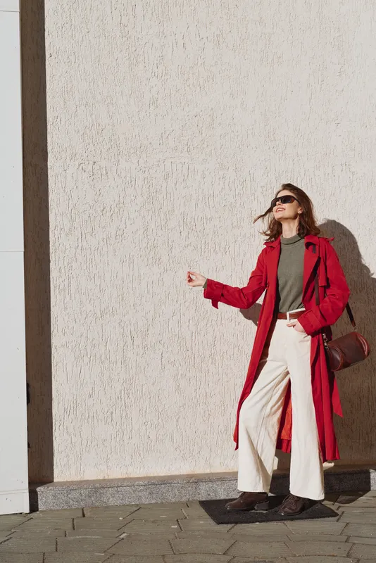 Stylish woman in red coat and green top stands beside a textured wall, sunglasses on, confident pose in daylight.