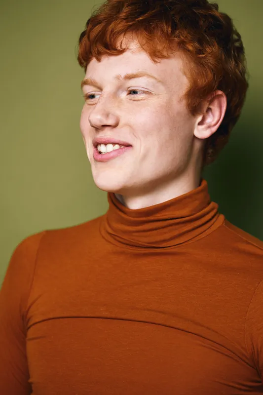 Young man with red hair wearing brown turtleneck sweater smiling and looking sideways with natural expression and green background in studio portrait.