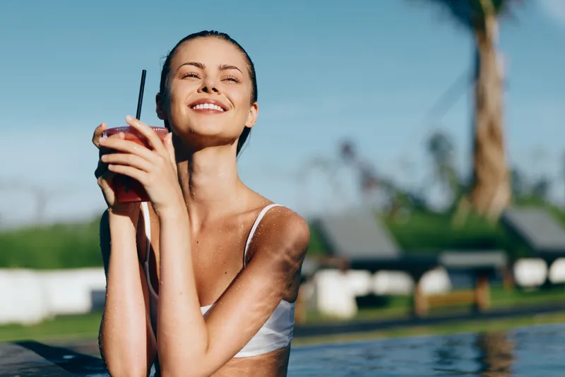A sunlit poolside moment shows a smiling woman holding a drink with a straw, wearing a white bikini top. Bright outdoor setting captures carefree summer lifestyle and refreshment.