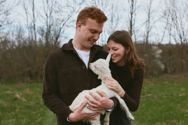 Loving couple outdoors, smiling softly while holding a goat kid, warm natural light, casual clothing, grassy field, spring atmosphere, gentle connection, candid lifestyle moment