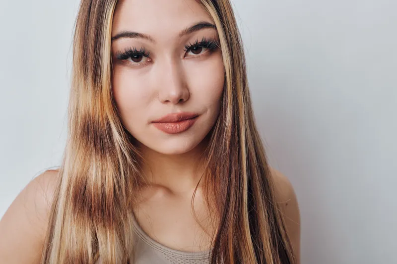 Young woman with long hair and natural makeup looking at camera with slight smile on neutral background. Portrait closeup with focus on face and eyes in soft lighting and casual style.