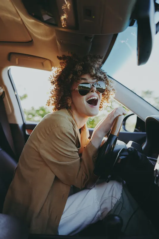 A joyful woman wearing sunglasses sits in a sunlit car, laughing while driving. A candid lifestyle moment with retro color tones and warm, cinematic glow.