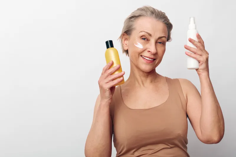 Mature woman with short grey hair applies face cream and holds two cosmetic bottles smiling on white background. Concept of skincare, beauty routine, anti aging and self care.