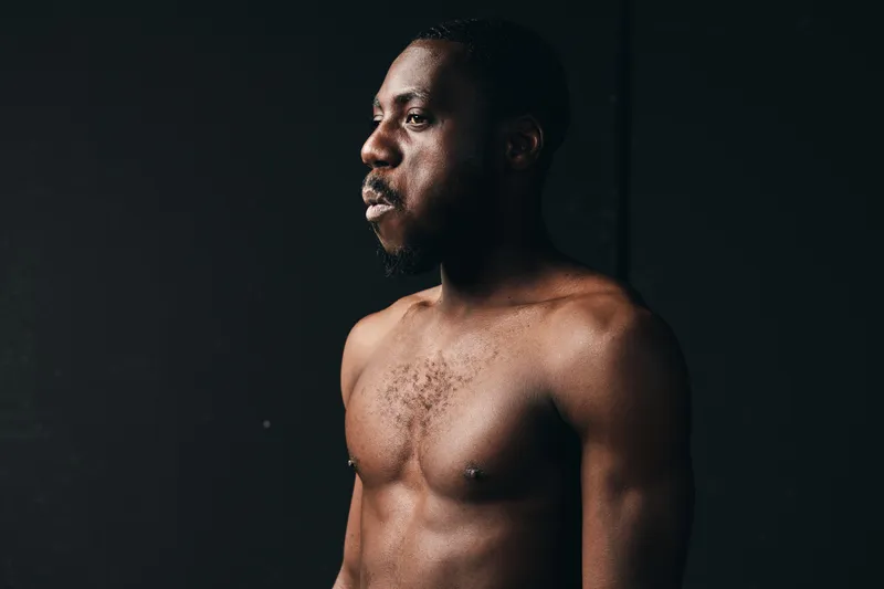 African man with dark skin and muscular body posing shirtless in creative conceptual visual style on black background. Portrait with dramatic lighting and serious expression in studio.