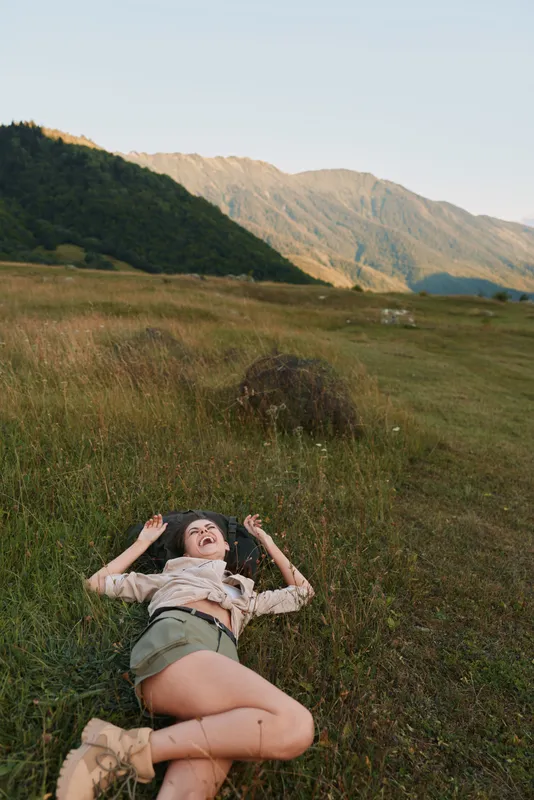 Young woman lying on grass field outdoors, enjoying nature, relaxed lifestyle, casual clothing, sunny day, mountain background, peaceful moment