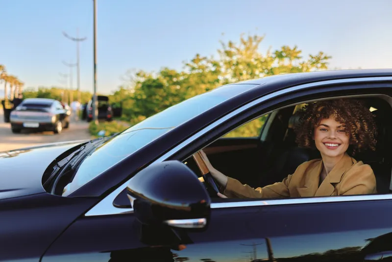 A smiling woman sits behind the wheel of a sleek car, looking out from the window with a carefree lifestyle vibe, enhanced by retro film color and warm, sunny tones.