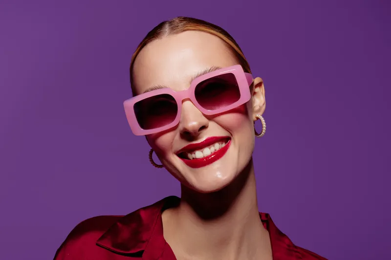 Smiling woman with pink sunglasses and red lipstick poses against purple background. Fashion portrait with bright colors, trendy style, and confident mood in studio lighting.