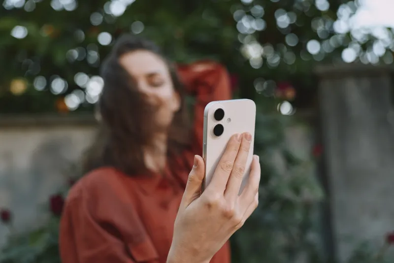 Young woman with long hair takes a casual selfie in a garden, smiling softly as she holds a smartphone up, with a blurred leafy background and warm natural light creating an inviting mood