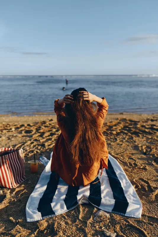 Young woman with long hair sits on a striped towel at the beach facing the calm sea while holding her hair with both hands in a relaxed pose during a sunny day.