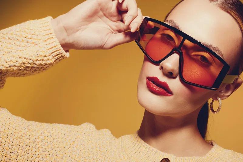 Stylish woman with red lipstick wearing oversized orange sunglasses and yellow sweater poses against orange background. Concept of fashion, beauty, accessories, and modern style.
