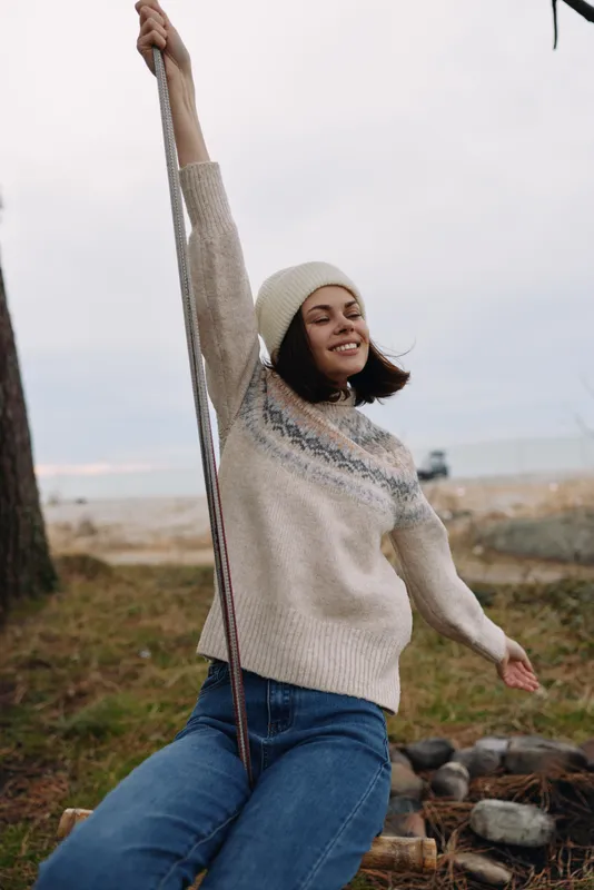 Woman swinging outdoors in a cozy sweater and beanie, by the coast with rocky shore. She smiles, enjoying a breezy day, casual fashion and relaxed mood in natural scenery.