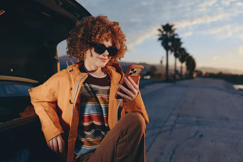 Woman lifestyle outdoors, wearing sunglasses and curly hair, seated by a car at sunset, relaxed smile while holding a smartphone, casual jacket, warm evening light, urban roadside background