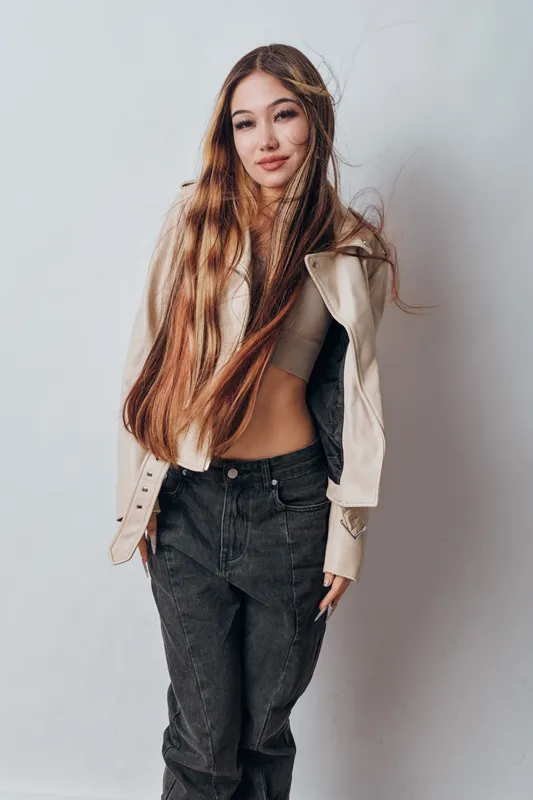 Young woman with long hair wearing cropped top and light jacket posing against white background. Casual fashion style with black pants and relaxed confident expression.