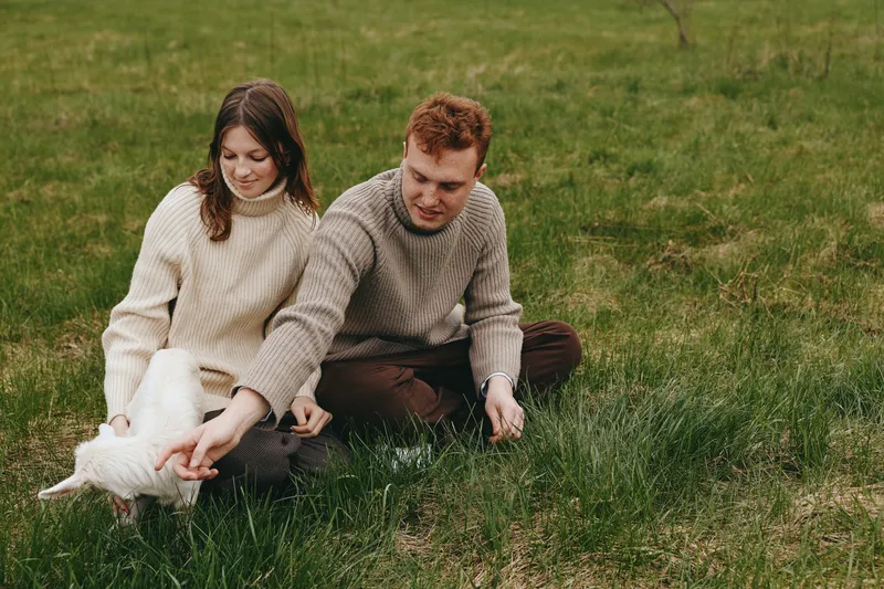 Couple sitting on grass stroking a baby goat kid in a sunny field, cozy sweaters, countryside lifestyle, connection with animals, peaceful moment, outdoor relaxation, soft natural lighting, warm and