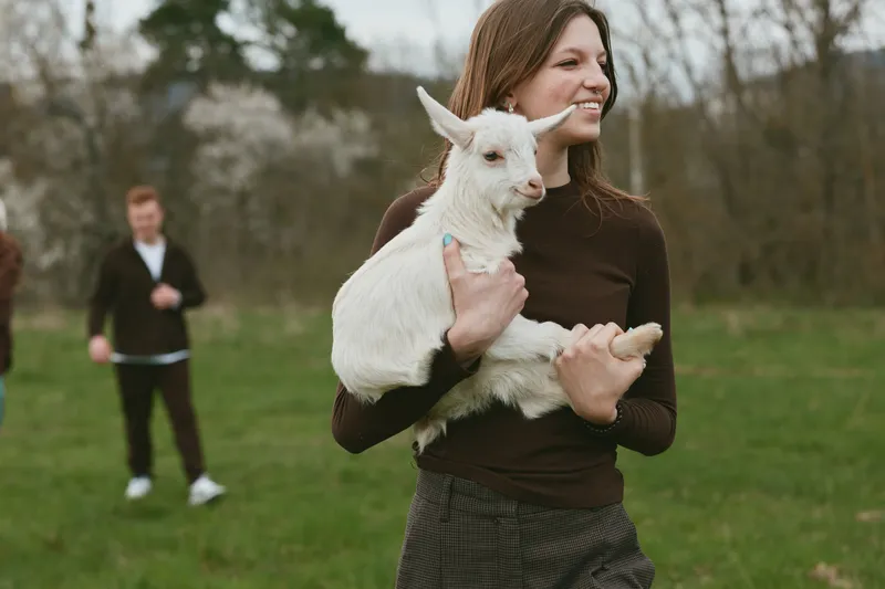 Lifestyle scene shows a smiling woman holding a white goat kid in a green park, with a man in the background, highlighting playful outdoor bonding and casual everyday joy for warm vibe.