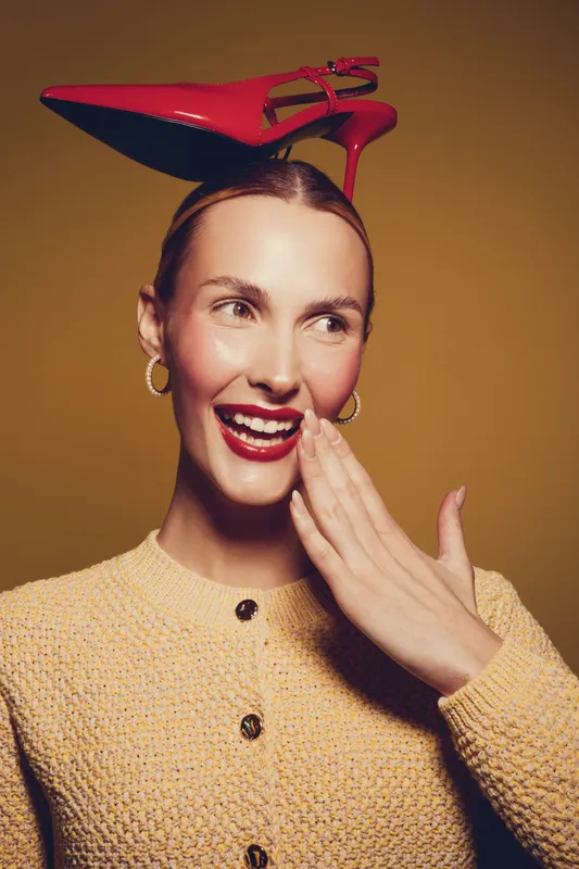 Smiling woman with red lipstick and hoop earrings wears red high heel shoes on her head. She covers her mouth with hand on brown background. Concept of fashion and playful mood.