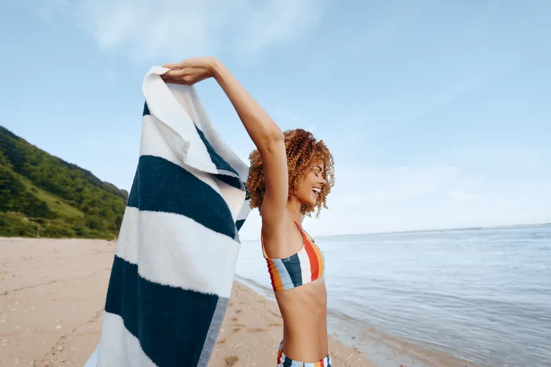 Sunlit beach scene shows a young woman with curly hair in a striped swimsuit lifting a striped towel above her head in a carefree, relaxed moment.