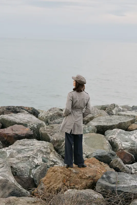Woman in trench coat and hat stands on rocky shore looking at calm sea horizon. Overcast sky with muted colors creates peaceful contemplative mood near water landscape.