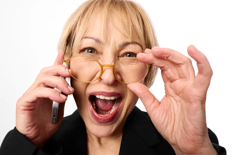 Woman with blonde hair, glasses held forward, joyful smile, speaking on phone, surprised open mouth, close portrait, white background, soft studio lighting, bright mood today