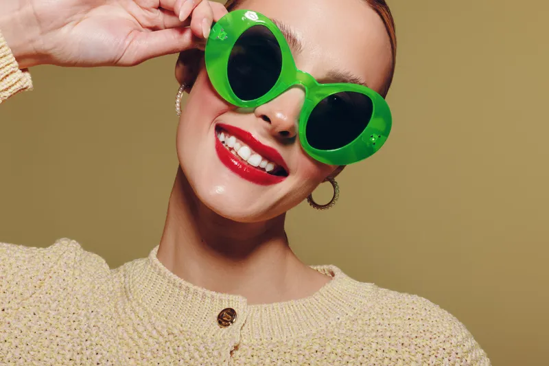 Smiling woman with bright red lipstick wears large green sunglasses and beige sweater. Close-up portrait on neutral background. Concept of fashion, style, and confidence.