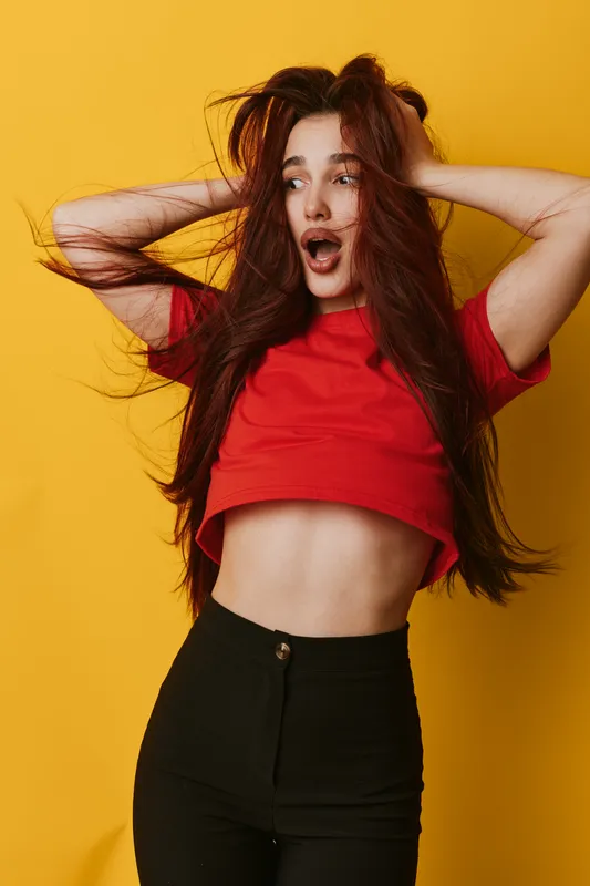 Woman with long hair in red shirt and black pants posing with hands in hair and surprised expression on yellow background. Fashion, emotion, beauty and casual style portrait.