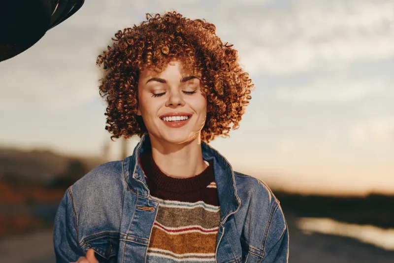 Smiling woman with curly hair in denim jacket and sweater enjoys golden hour outdoors at sunset, eyes closed and relaxed posture, lifestyle concept of happiness and confidence