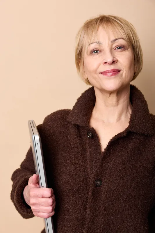 Woman mature smiling portrait holding tablet, beige studio background, dark jacket, soft studio lighting, calm expression, wellness concept for health and self care