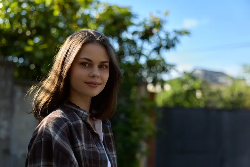 Young woman with shoulder length brunette wearing a plaid shirt stands outdoors with a calm smile in a sunlit garden, conveying a casual lifestyle moment and natural confidence