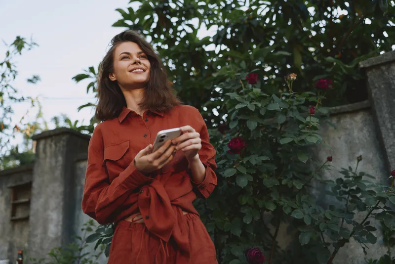 Smiling woman in red dress outdoors, holding a smartphone. Natural light highlights a garden backdrop, creating a lifestyle moment of casual chic and self expression
