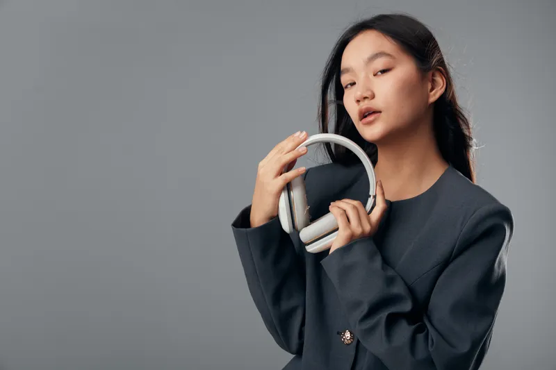 Woman holding headphones posing on gray background with confident expression wearing dark blazer. Music equipment, technology and fashion concept with stylish female and audio device.